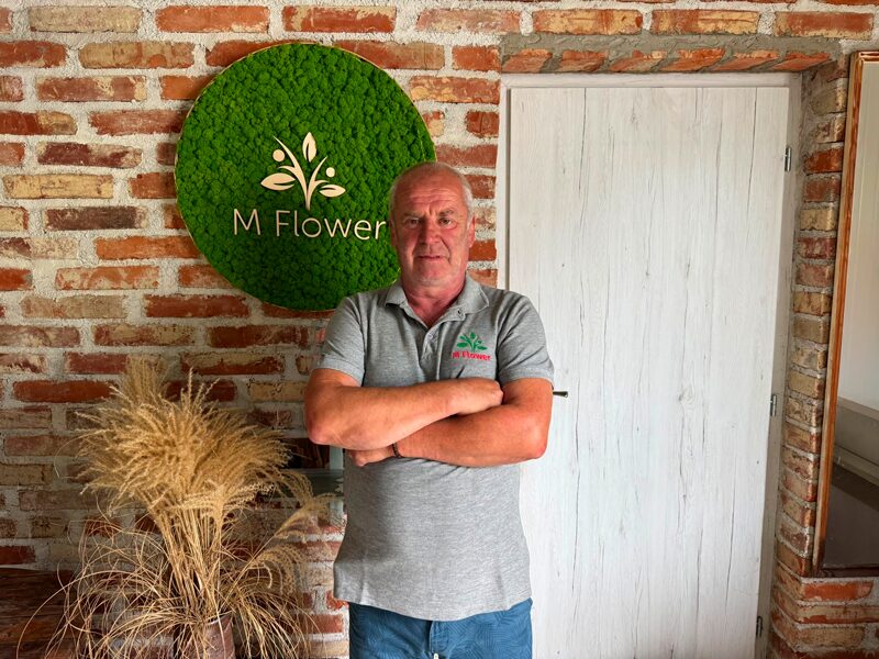 Alternatívny text: Spoluzakladateľ firmy M Flower v interiéri pred zeleným logom M Flower a tehlovou stenou